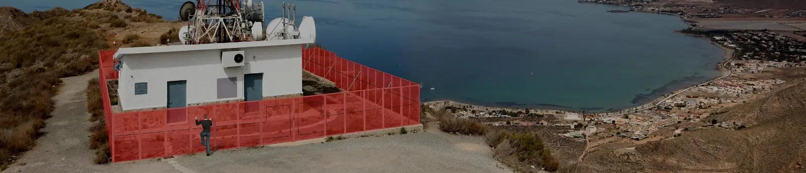 Visualisierung: Person klettert am Zaun um eine Funkstation auf einer Anhöhe über der Küste; der Zaun ist rot als Alarmzone markiert – Visualisierung Southwest Microwave (Symbolbild): Zaundetektion am Gelände.